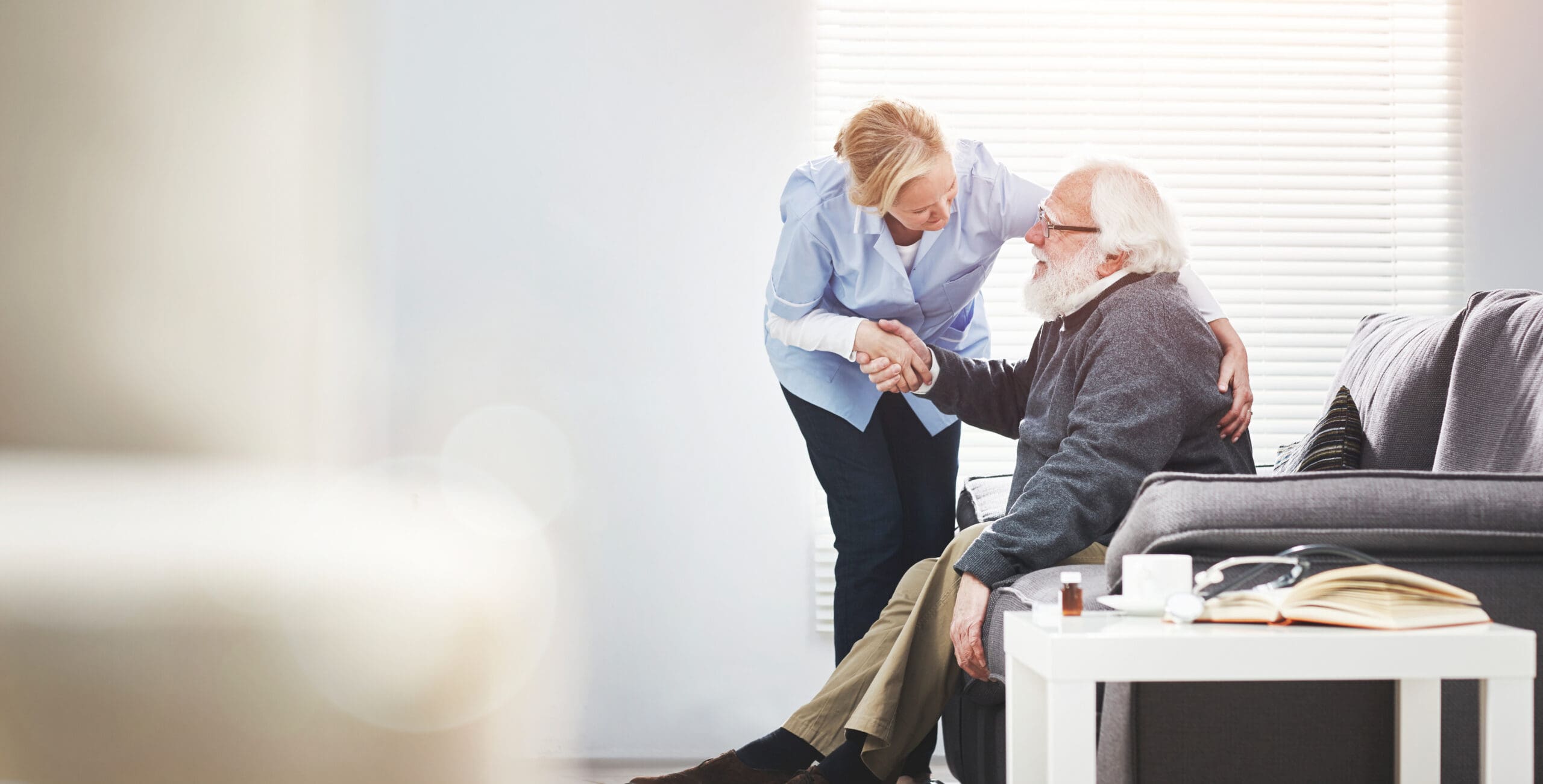 A healthcare worker leaning over and shaking hands with an elderly man sitting on a gray couch in a bright room with blinds. The woman has blonde hair and is wearing a light blue uniform, while the elderly man has white hair, a beard, glasses, and is dressed in a dark sweater and beige pants. There is a small white table beside him with medication bottles, a notebook, and a cup. The scene conveys a caring and supportive interaction.