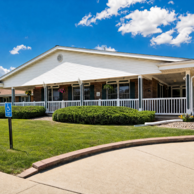 A single-story building with a white exterior and a large front porch, surrounded by neatly trimmed bushes and green grass, under a bright blue sky with scattered clouds. There is a handicapped parking sign in the foreground.