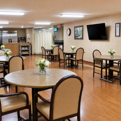 A spacious dining area with round tables decorated with white flower arrangements, beige cushioned chairs, a wall-mounted TV, a clock, framed artwork, and a kitchenette in the background.