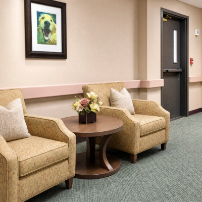 A waiting area with two beige armchairs with pillows, a round wooden table with a flower arrangement, a framed dog portrait on the wall, and a door on the right side.