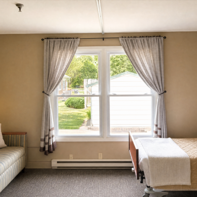 A large window with beige curtains tied back, revealing a view of a green lawn, trees, and neighboring houses outside. The room has a beige wall and a carpeted floor. Part of a striped sofa is visible on the left, and a bed with a white blanket is on the right.