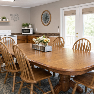 A dining area with a large wooden table surrounded by six matching wooden chairs. The table has a rectangular metal container with a bouquet of white and yellow flowers. In the background, there is a kitchen counter with a microwave, potted plants, and wall decor including a round clock and a sign that reads 
