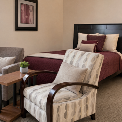 A cozy hotel room features a bed with a dark headboard, maroon and beige pillows, and matching maroon bedding. In front of the bed is a patterned armchair with wooden arms, and beside it a small wooden side table with a potted plant. A framed abstract artwork hangs on the wall above the sofa.