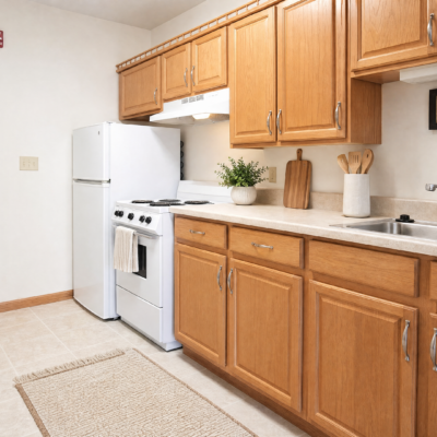 Kitchen with wooden cabinets, white stove, white refrigerator, and beige countertop. Decor includes a potted plant, wooden cutting boards, and a white utensil holder. Rug on the light-colored tiled floor.