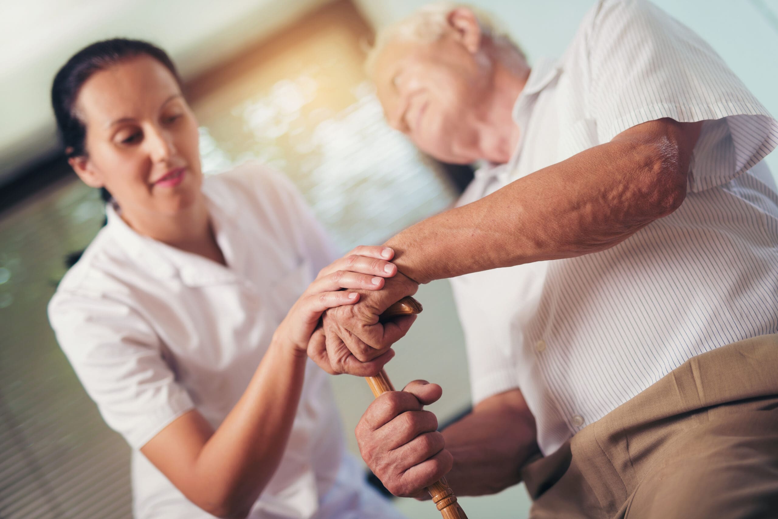 A caregiver assisting an elderly man by holding his hand and providing support with a walking cane in a healthcare setting.